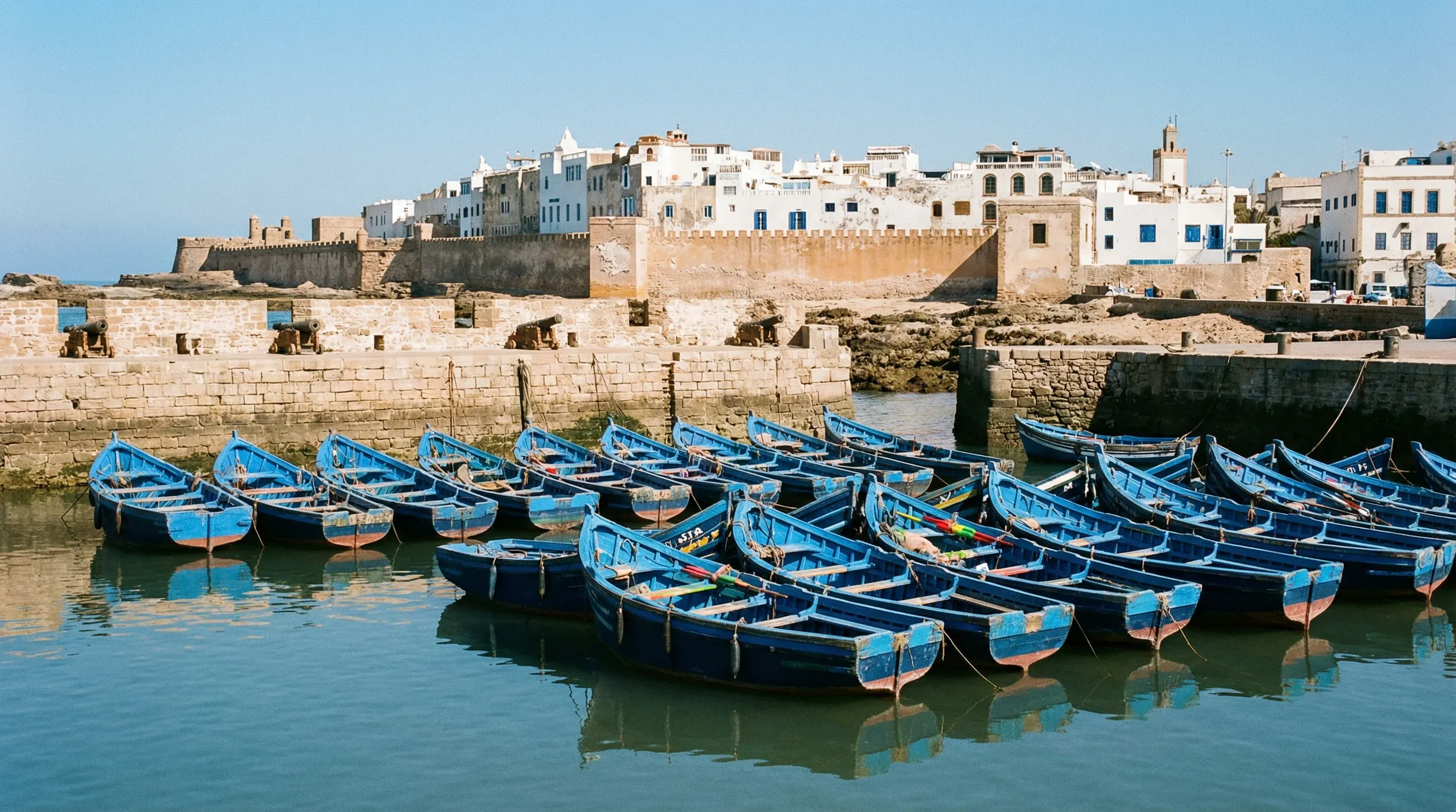 Bright blue wooden fishing boats sit in the harbor of Essaouira with the historic stone sea walls and white city buildings in the distance.