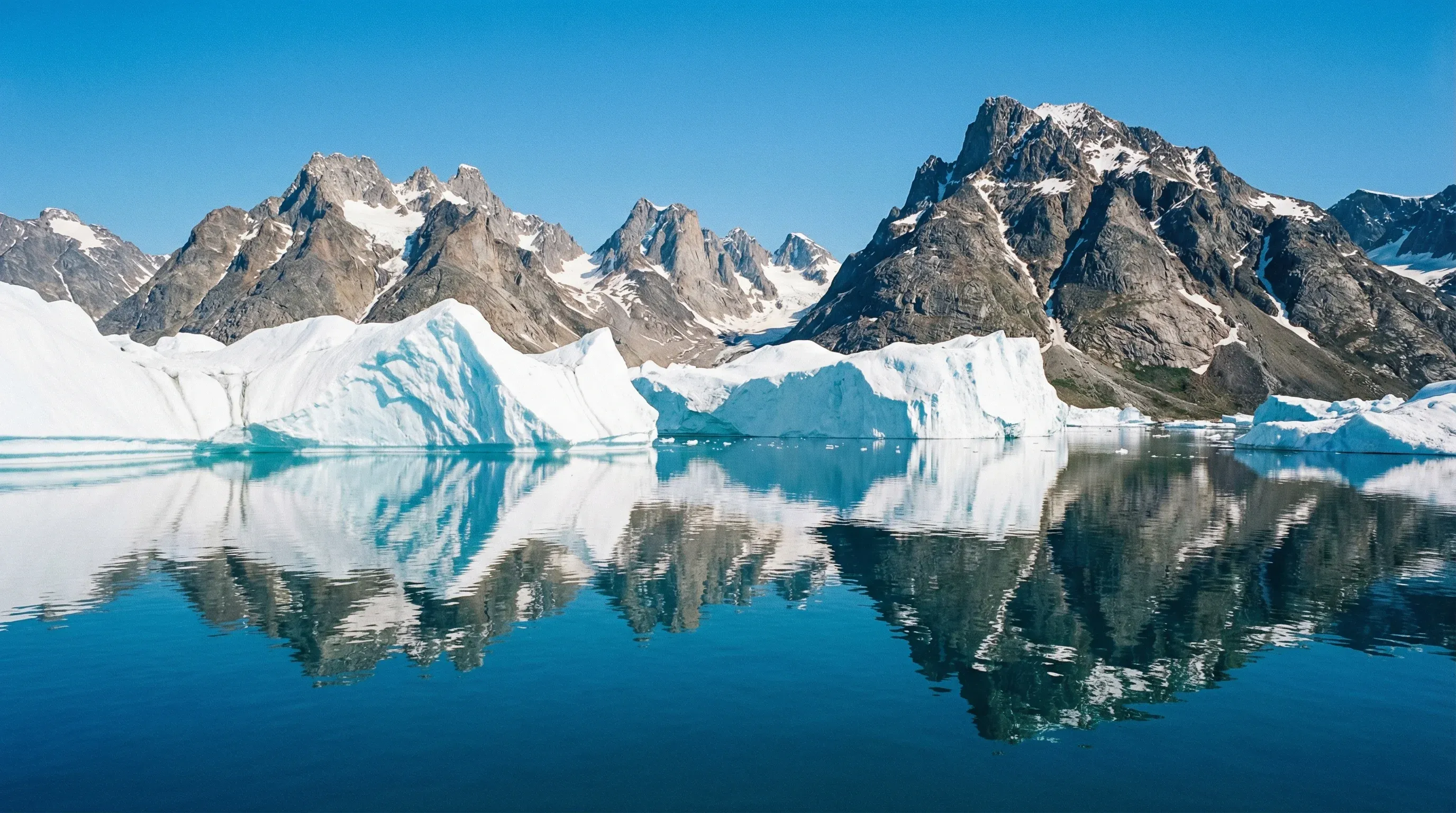Large icebergs floating in the blue waters of Sermilik Fjord, East Greenland, with sharp, snow-dusted mountains in the background under a clear sky.