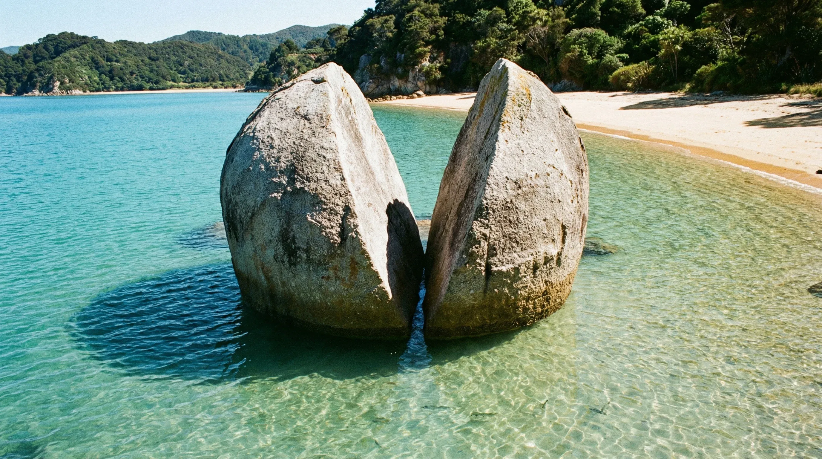 A large circular granite rock split down the middle sits in clear turquoise water near a sandy beach.