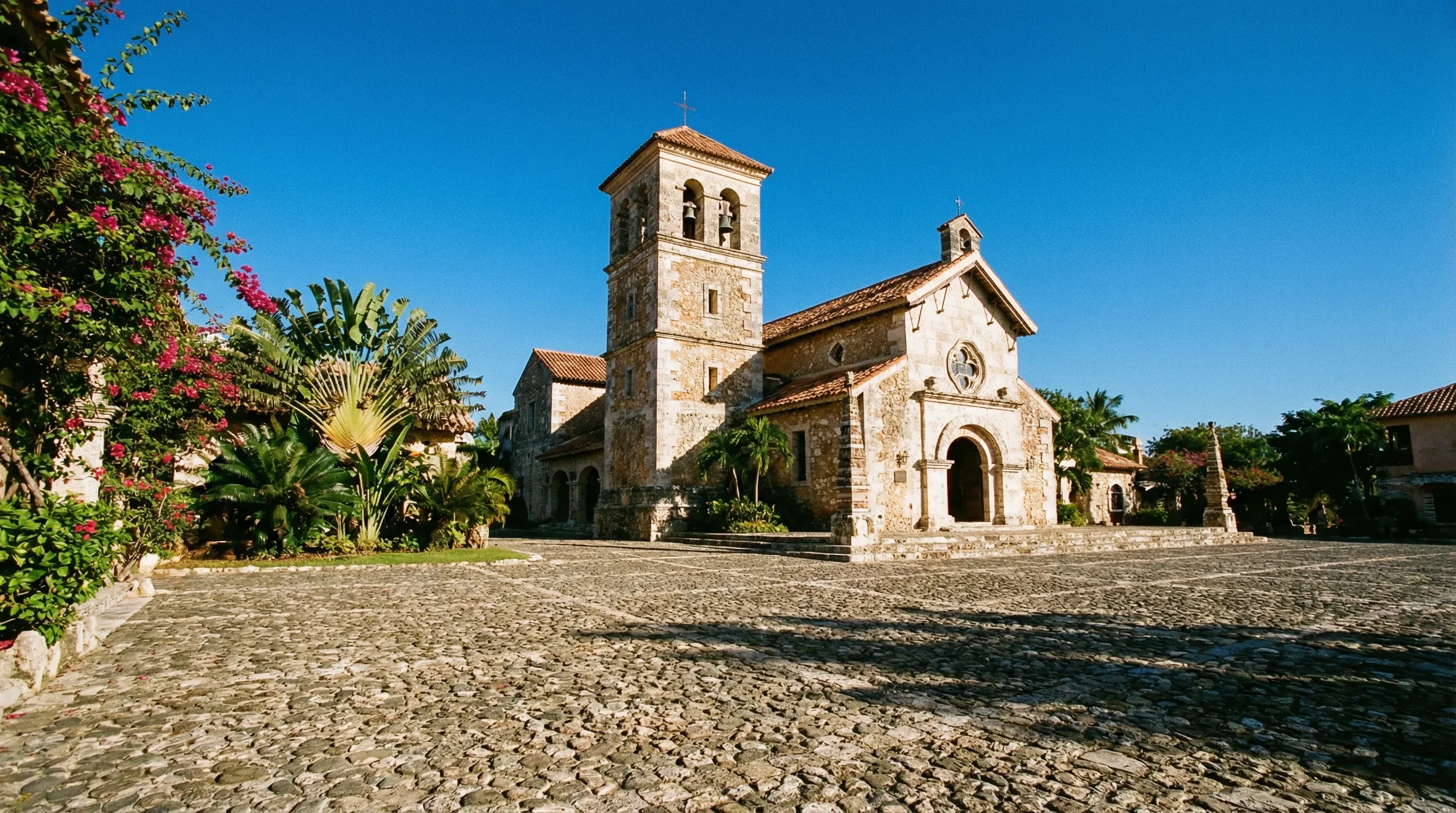 The coral stone St. Stanislaus Church and cobblestone plaza in Altos de Chavón, La Romana, under a clear blue sky.