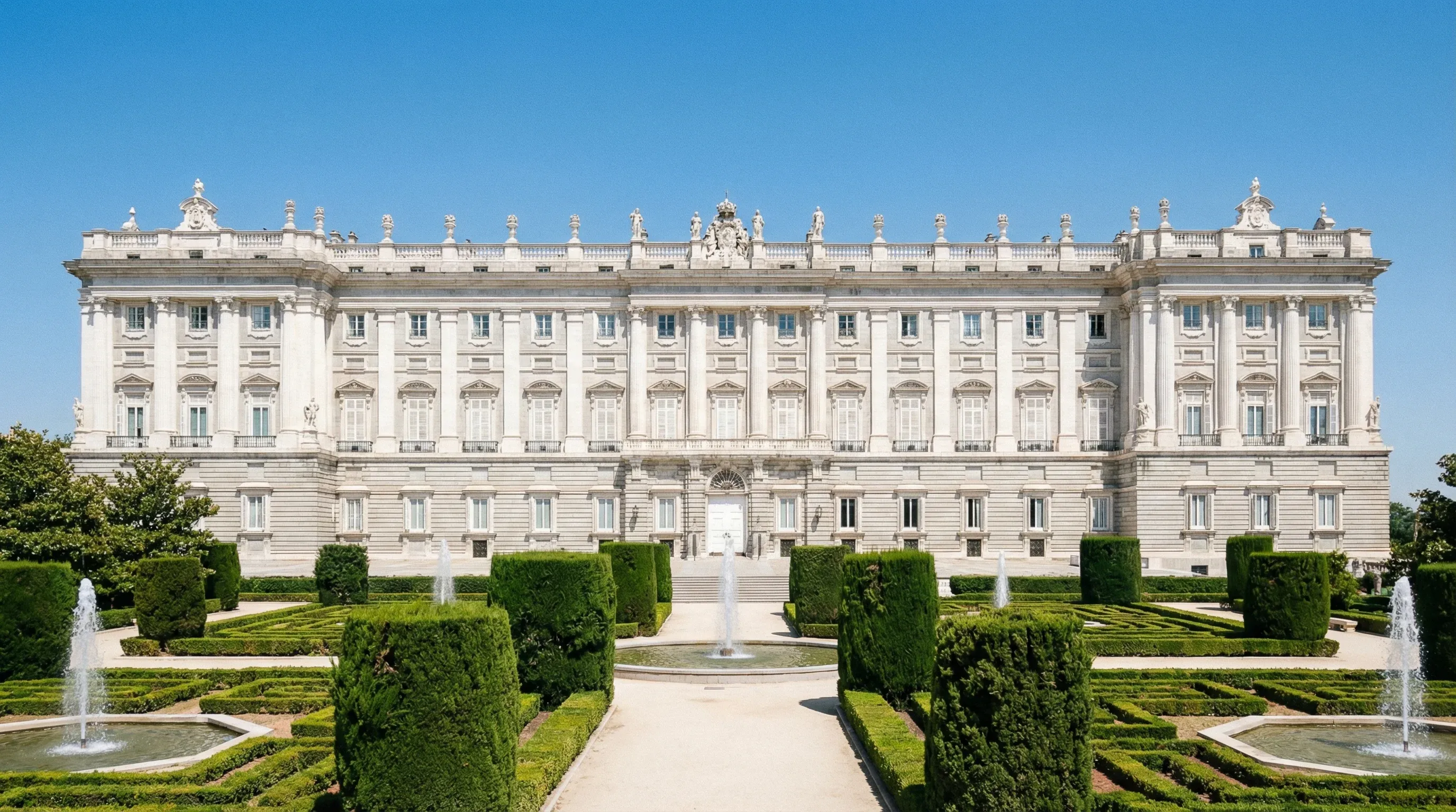 The Royal Palace of Madrid viewed from the Sabatini Gardens with its white stone facade under a bright sky.
