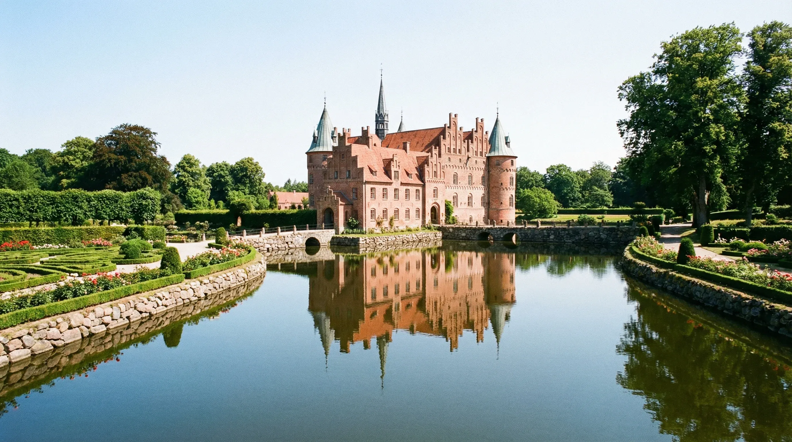 A red brick Renaissance castle with pointed spires reflected in a water moat, surrounded by green gardens.