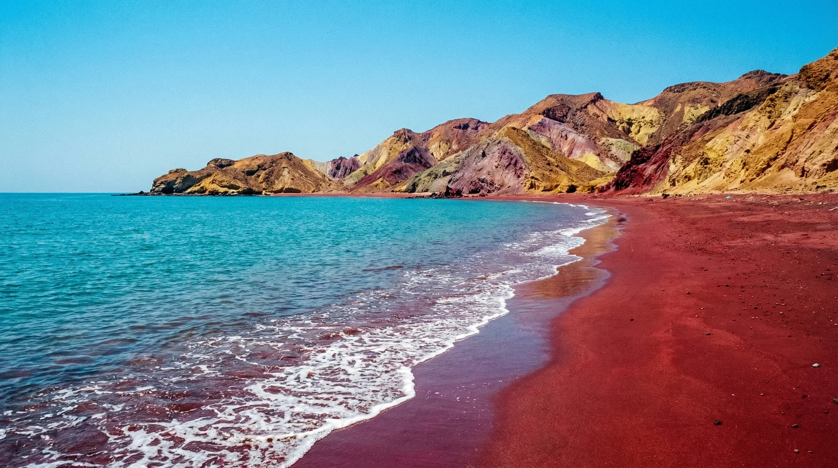 A shoreline view showing the meeting point of bright red sand and turquoise ocean water on Hormuz Island.