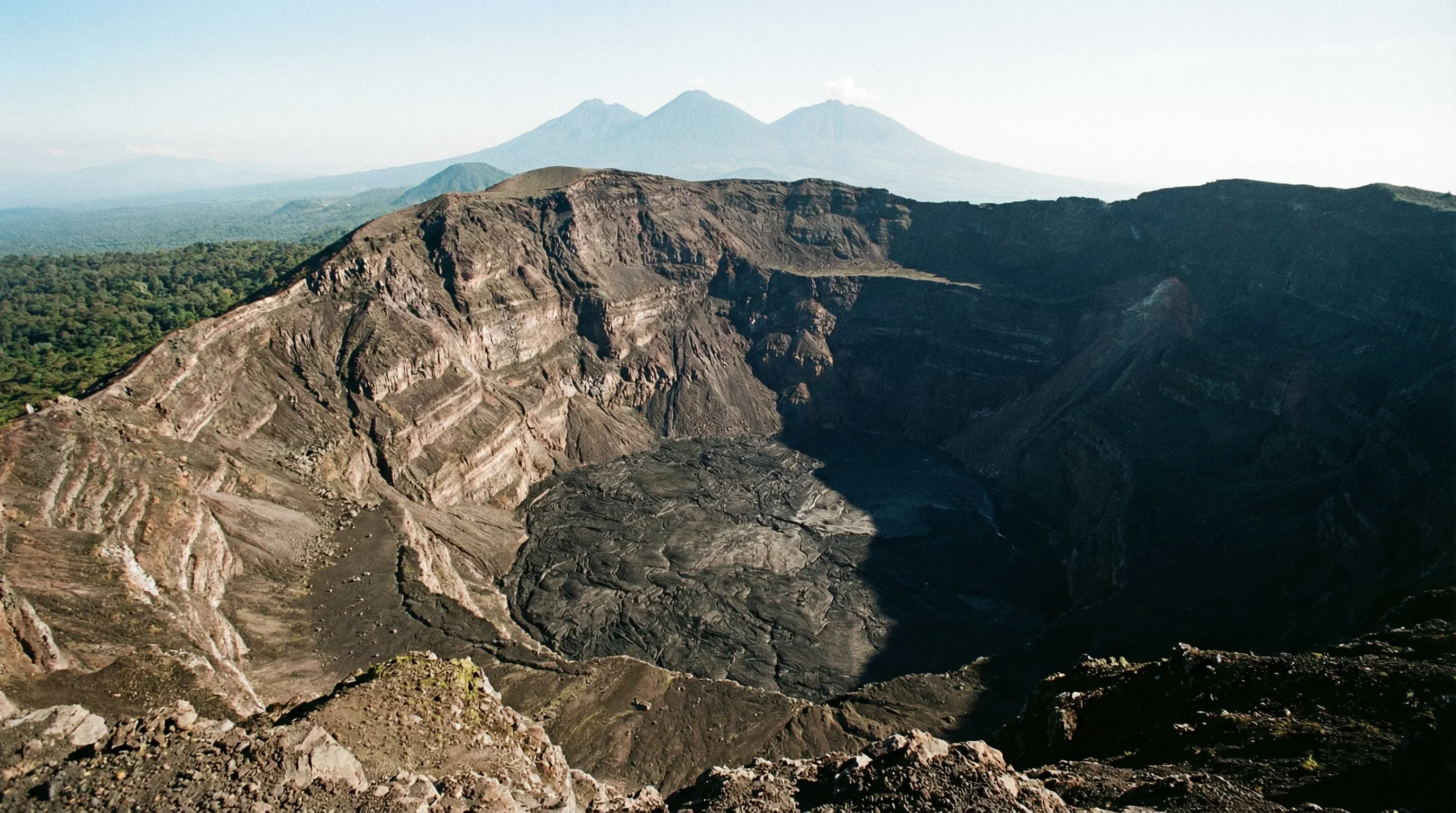 The wide volcanic crater of Mount Nyiragongo with steep rocky walls and other mountain peaks in the distance.