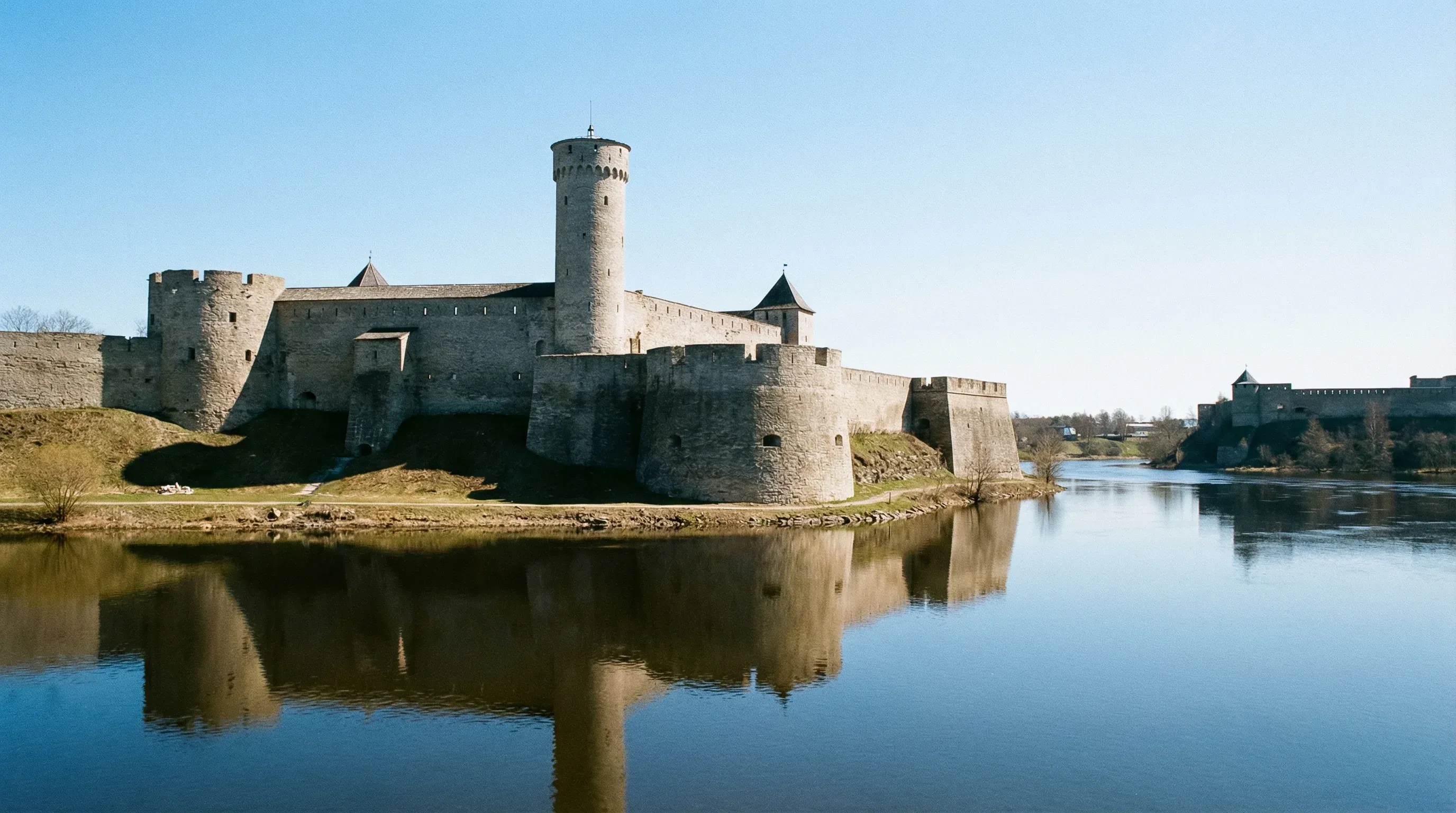 The medieval Narva Castle and its tall stone tower overlooking the Narva River under a blue sky.