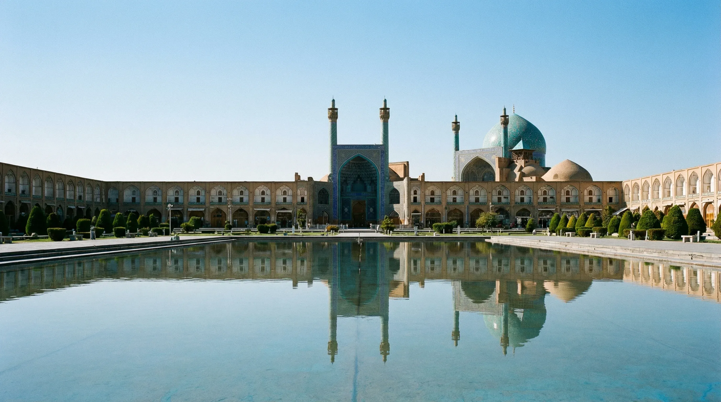 A wide view of the Shah Mosque with its blue-tiled dome and minarets reflected in a large pool at Naqsh-e Jahan Square in Isfahan.