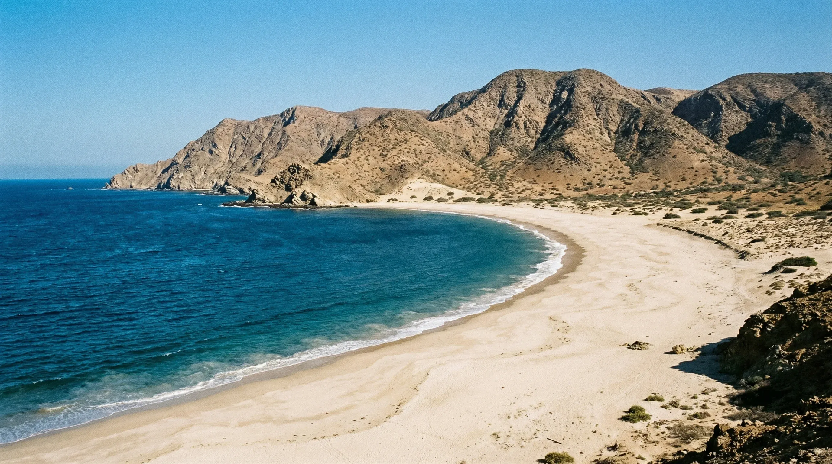 A wide view of the crescent-shaped Baía Azul beach with blue ocean water and dry coastal hills in Benguela, Angola.