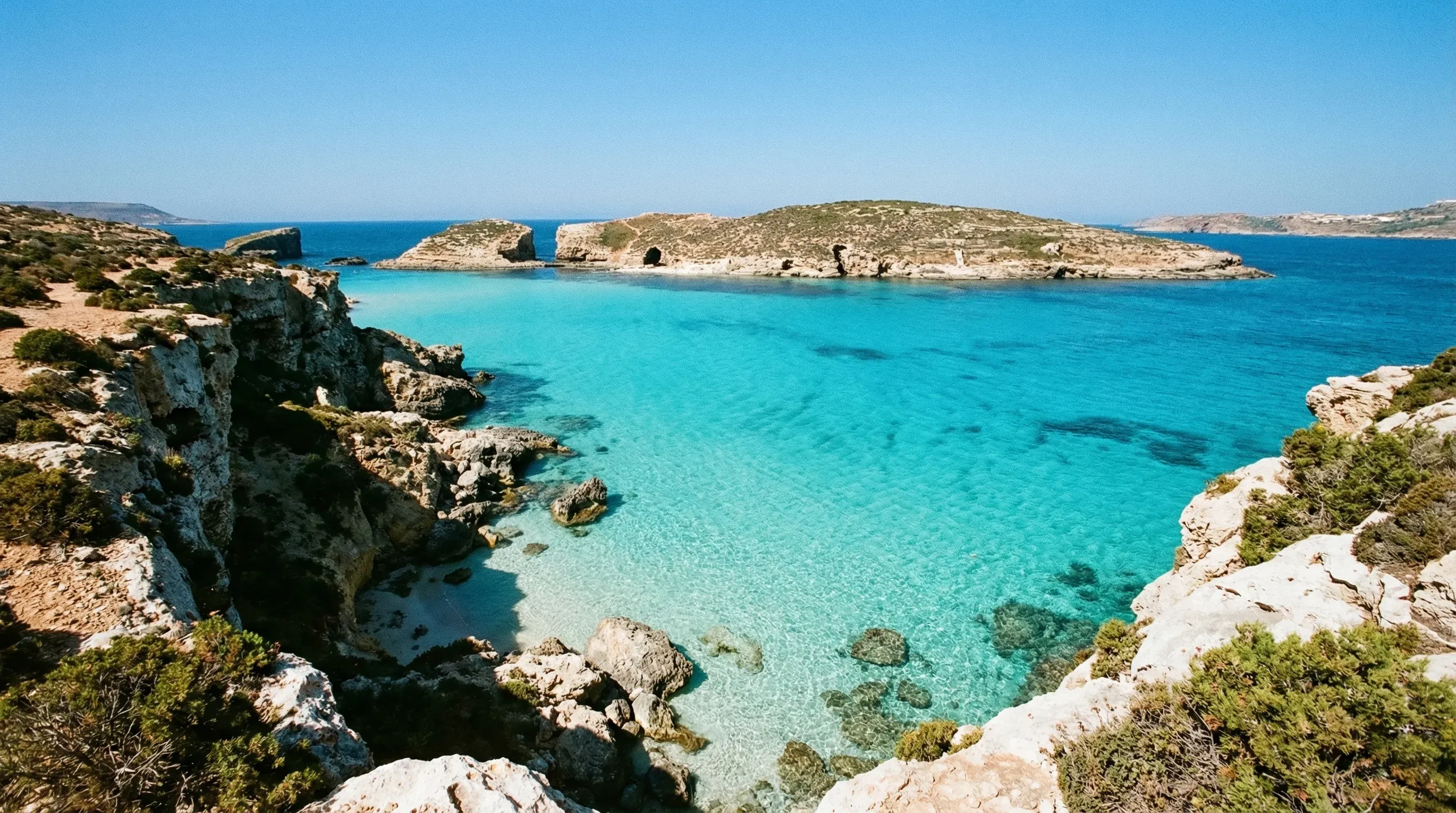 A high-angle view of the turquoise waters of the Blue Lagoon in Comino, showing the rocky coastline and the small islet of Cominotto under a clear sky.