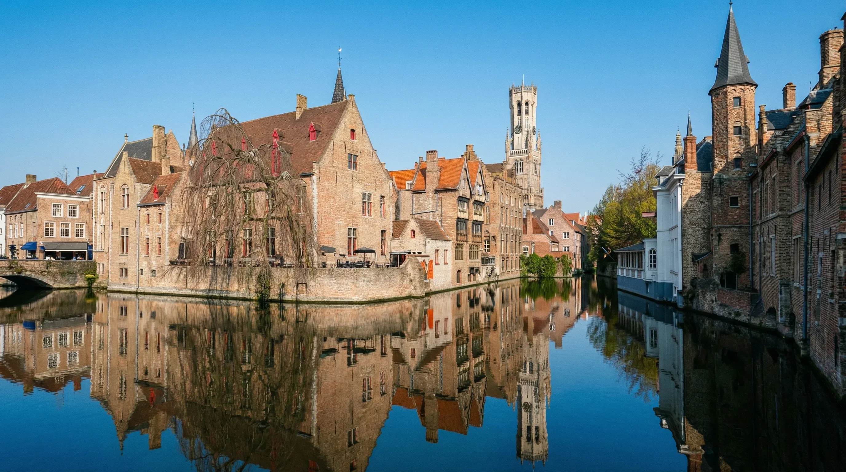 A view of the Rozenhoedkaai canal in Bruges, featuring historic brick buildings and the Belfry tower under a bright midday sun.