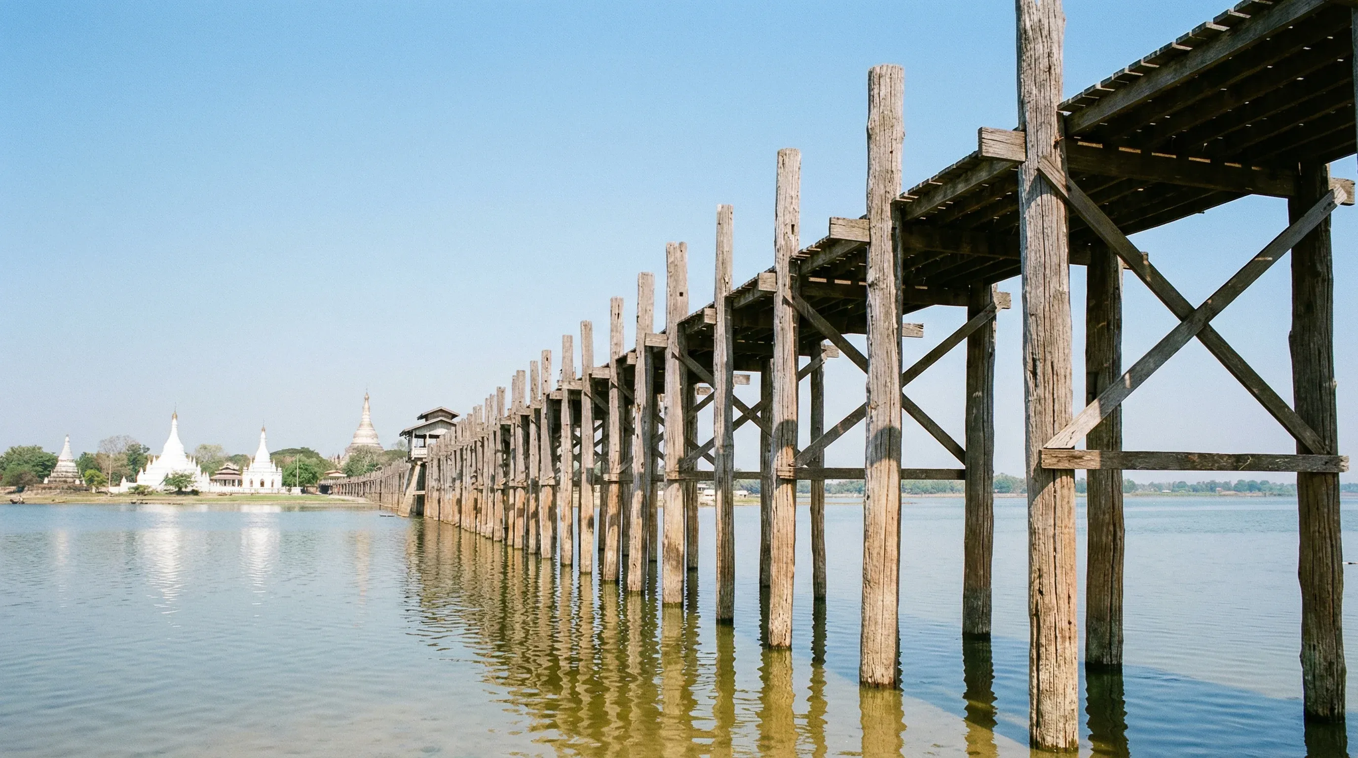 The long, historic teak U Bein Bridge spanning Taungthaman Lake under a bright midday sun.