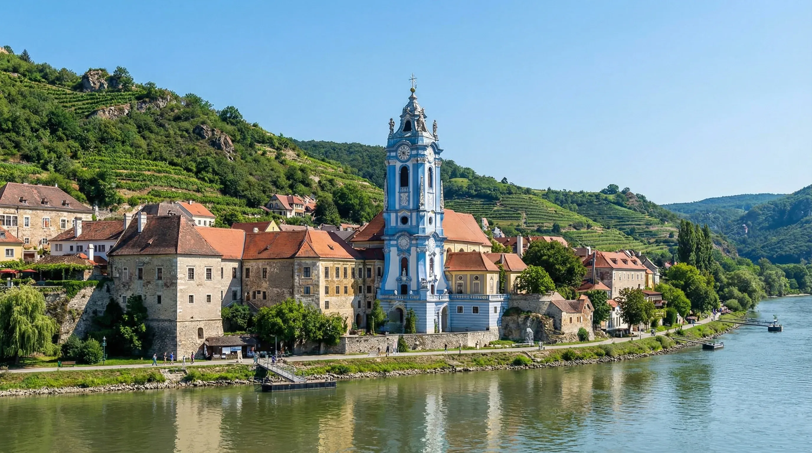 The blue baroque tower of Dürnstein Abbey stands prominently beside the Danube River, with terraced vineyards on the hills behind.