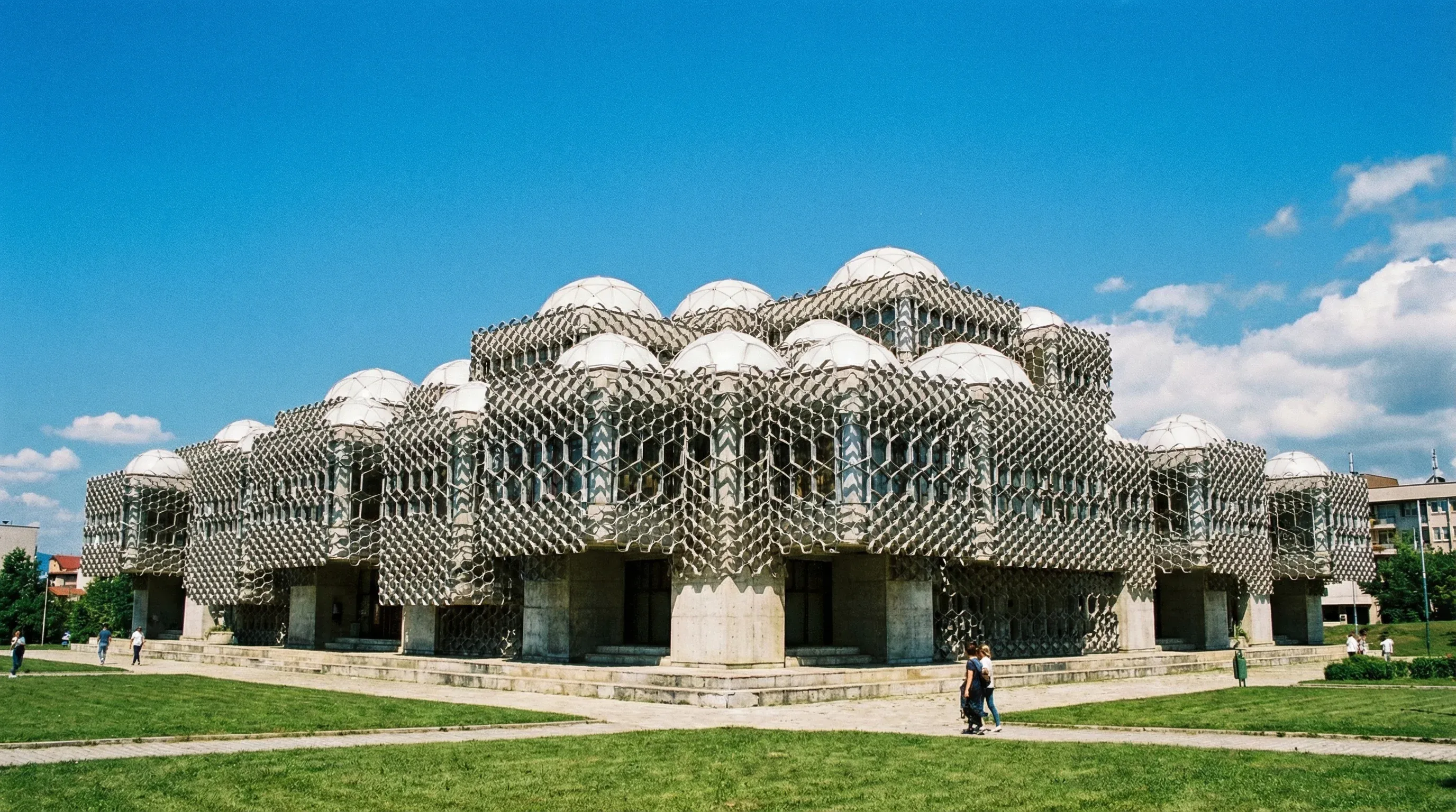 The National Library of Kosovo in Pristina, showcasing its unique metal grid exterior and white domes against a bright blue sky.