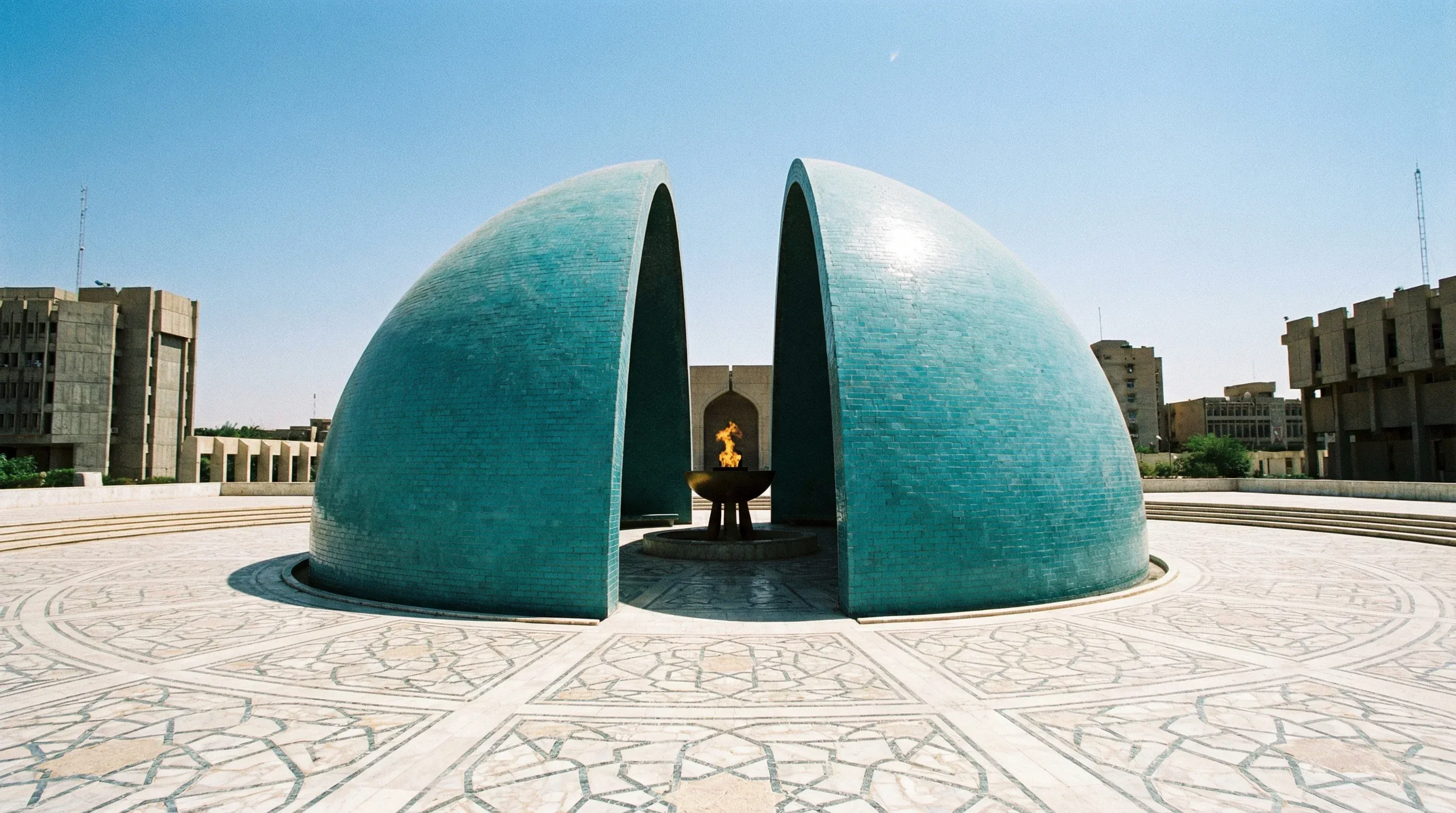 A split turquoise dome monument on a large circular platform in Baghdad under a clear sky.