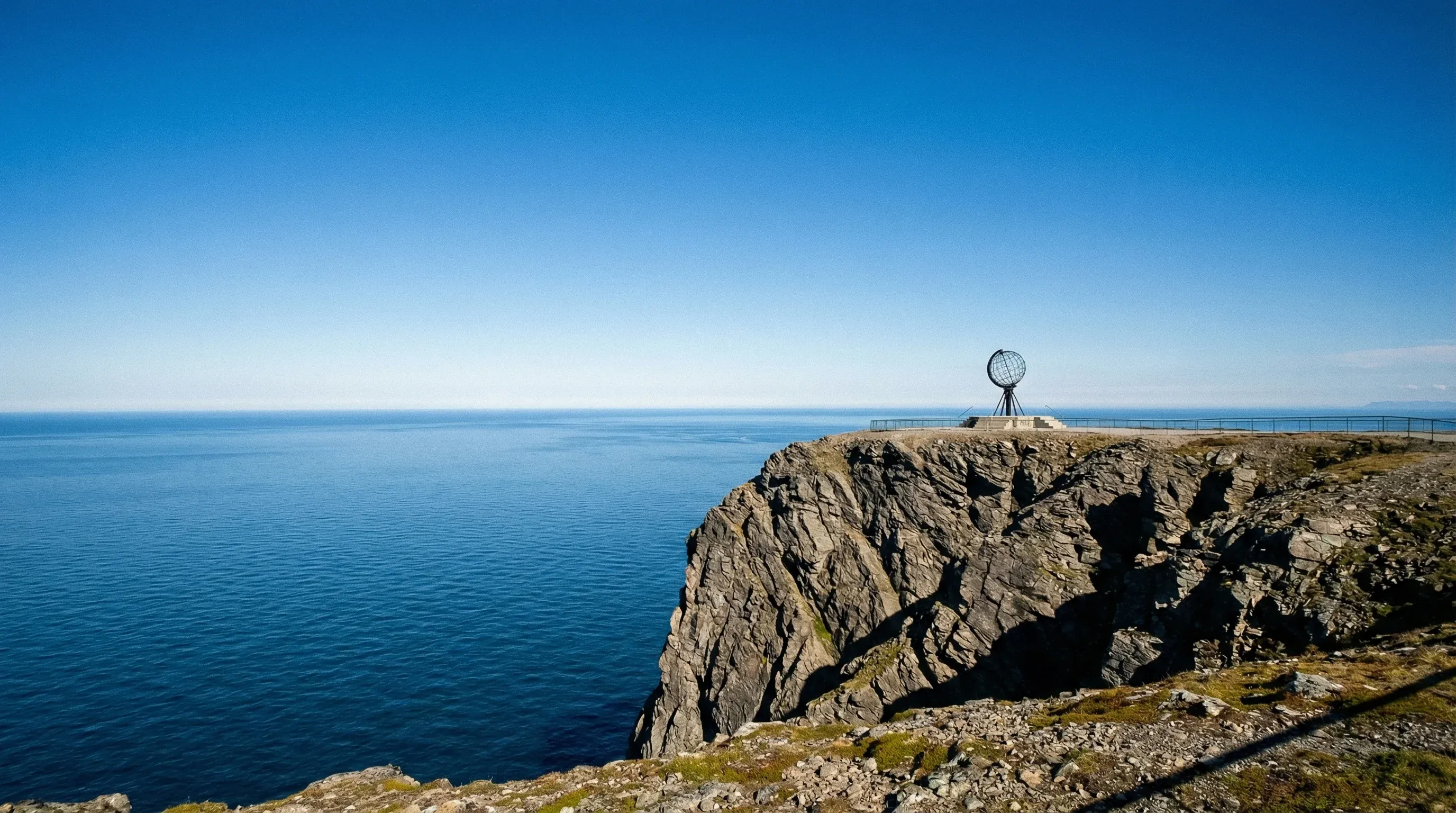 The iron globe monument on the edge of the high North Cape cliffs overlooking the Arctic Ocean under a clear sky.