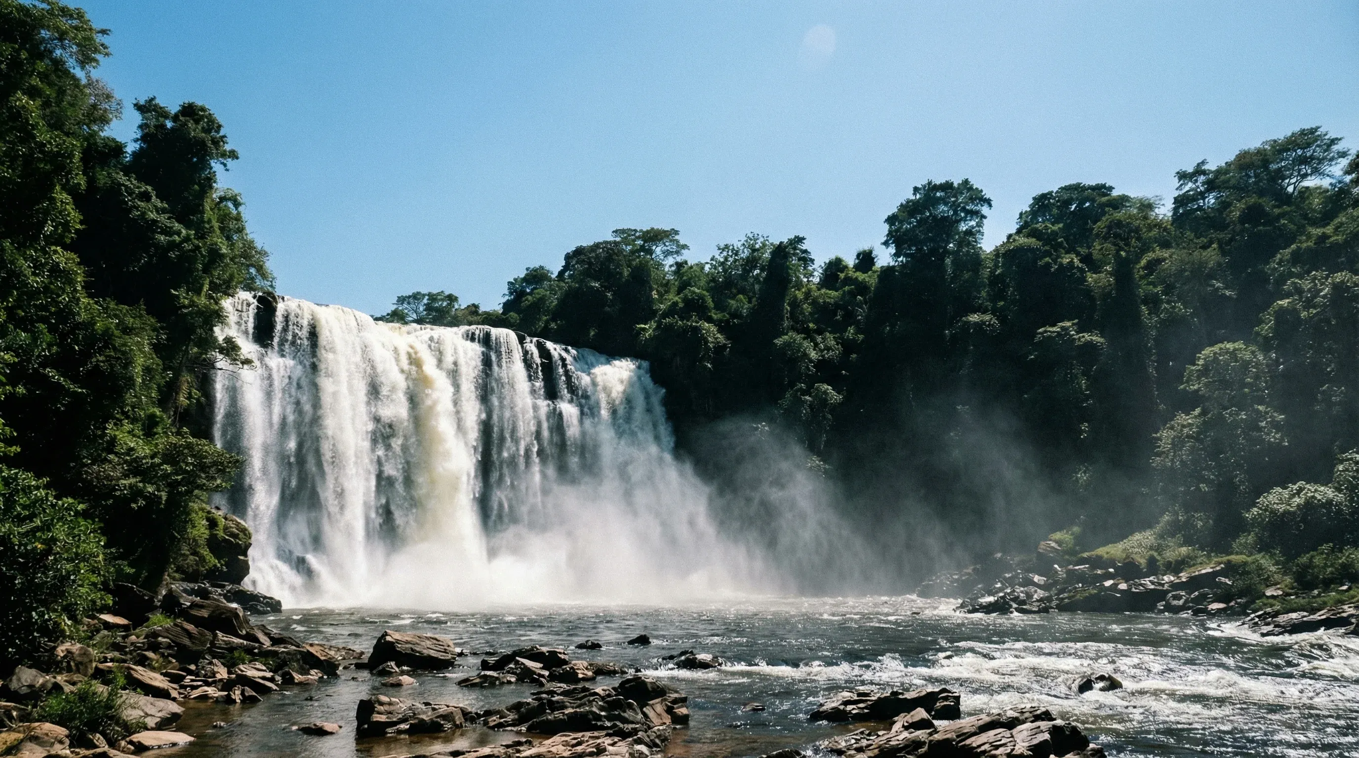 Lumangwe Falls in Northern Zambia, showing a wide curtain of water cascading over a rocky ledge into a mist-filled basin surrounded by forest.