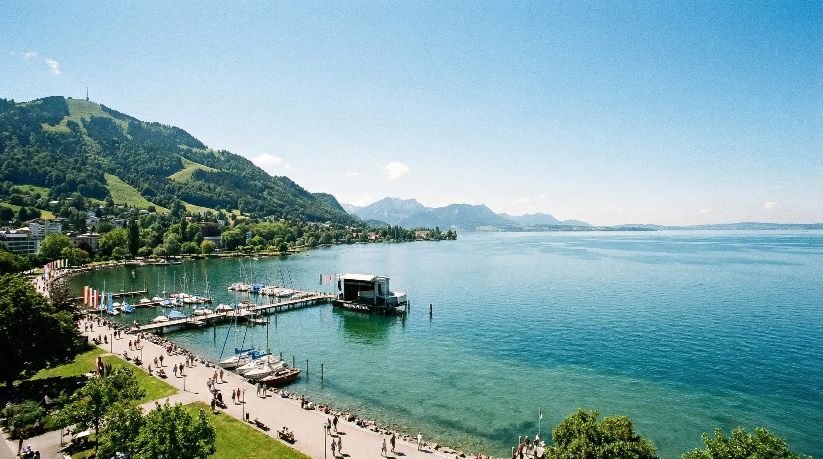 A wide view of the expanse of Lake Constance from the shore at Bregenz, with green mountains in the background under a clear sky.