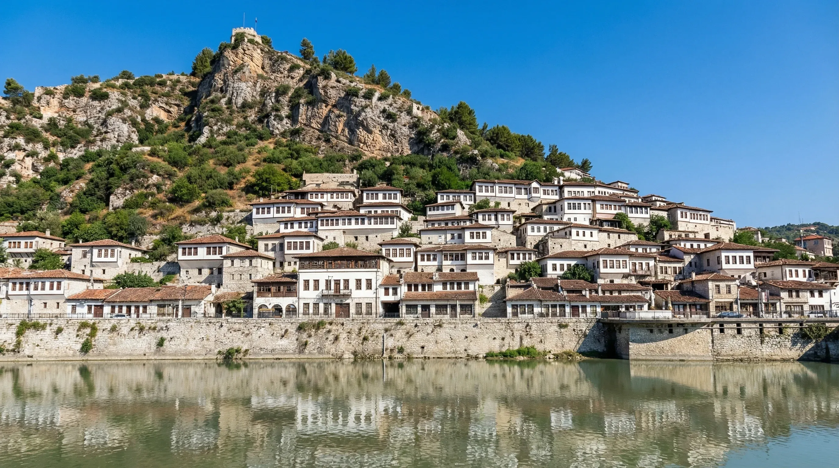 Rows of historic Ottoman-era white houses with many windows on a hillside in Berat, seen from across the Osum River.