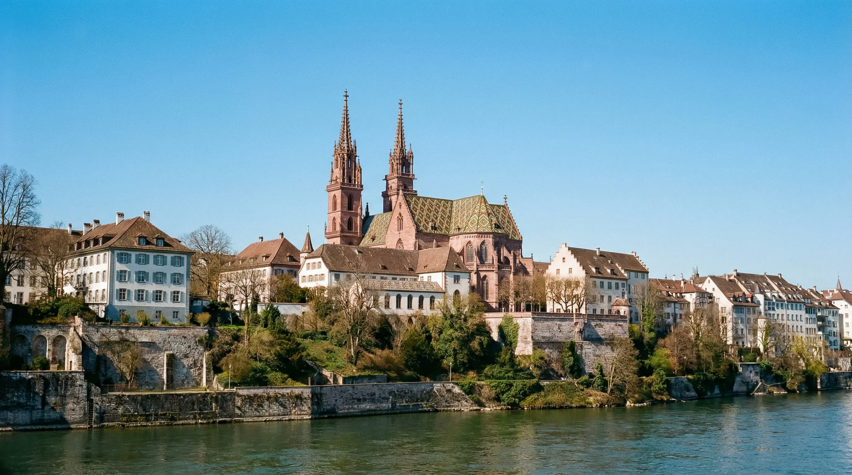 A wide-angle view of the red sandstone Basel Minster overlooking the Rhine River under a clear sky.