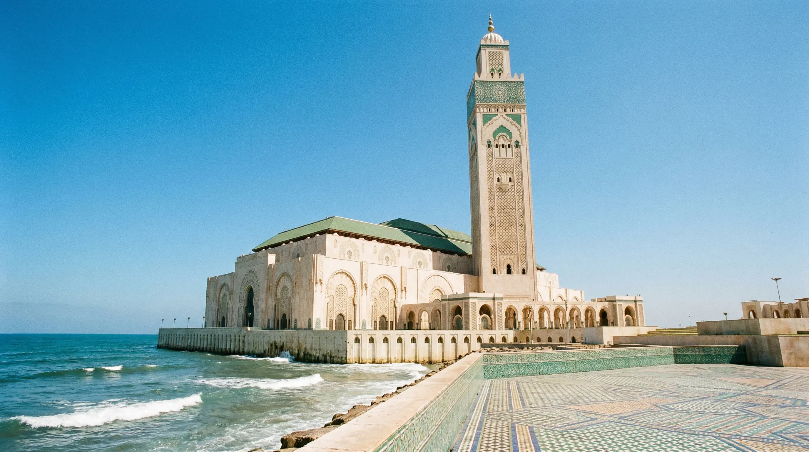 The massive Hassan II Mosque with its tall minaret stands on the Casablanca coastline next to the Atlantic Ocean under a clear blue sky.