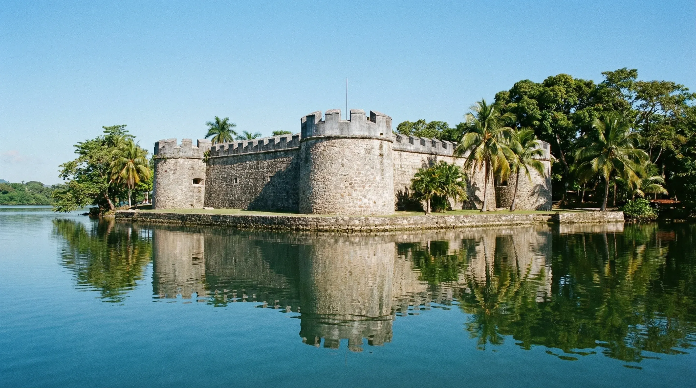 A historic stone fortress with circular towers sitting on the edge of the Río Dulce river.