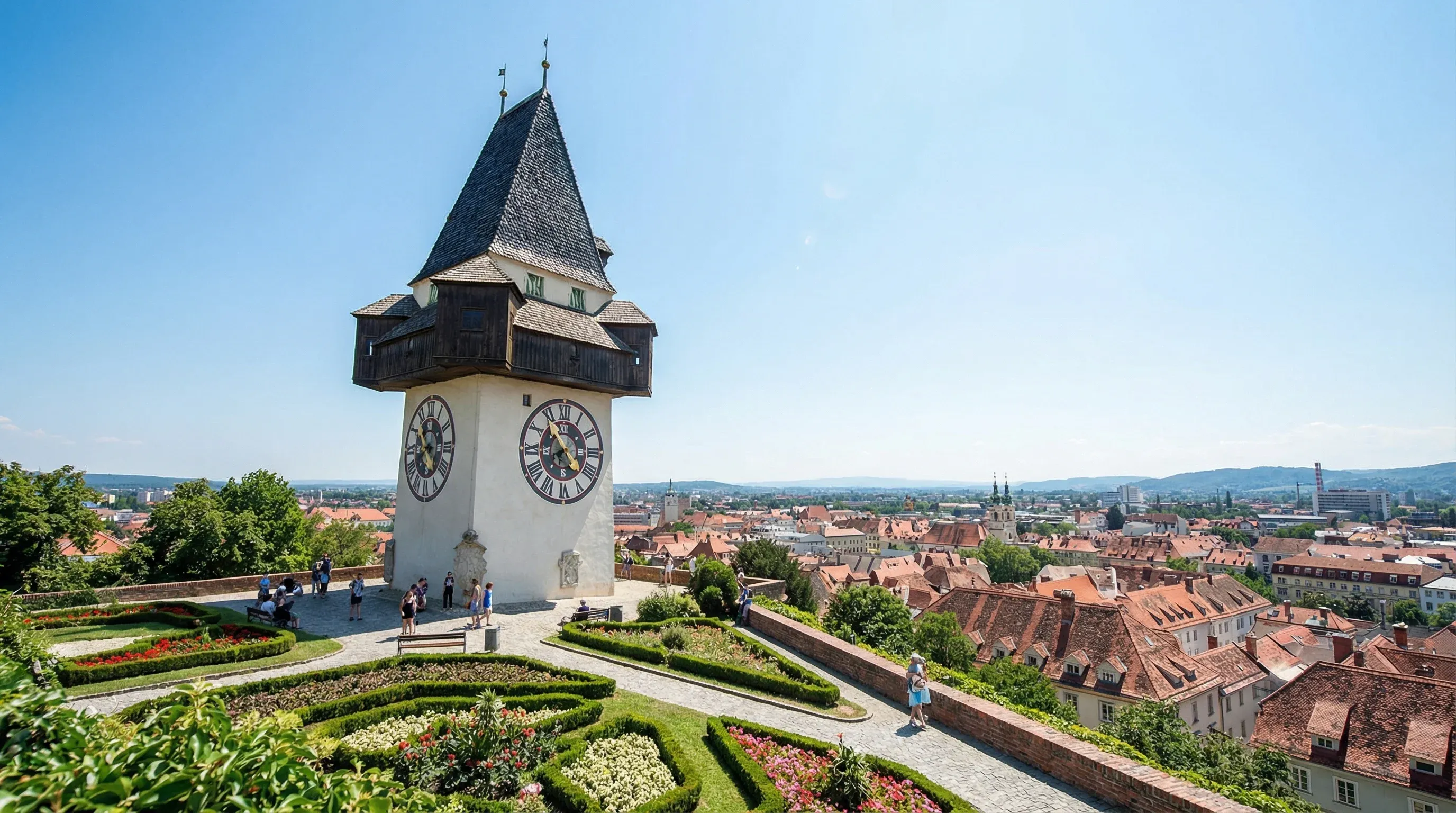 The historic Grazer Uhrturm clock tower stands on a hill overlooking the red-tiled roofs of the Graz old town under a clear sky.