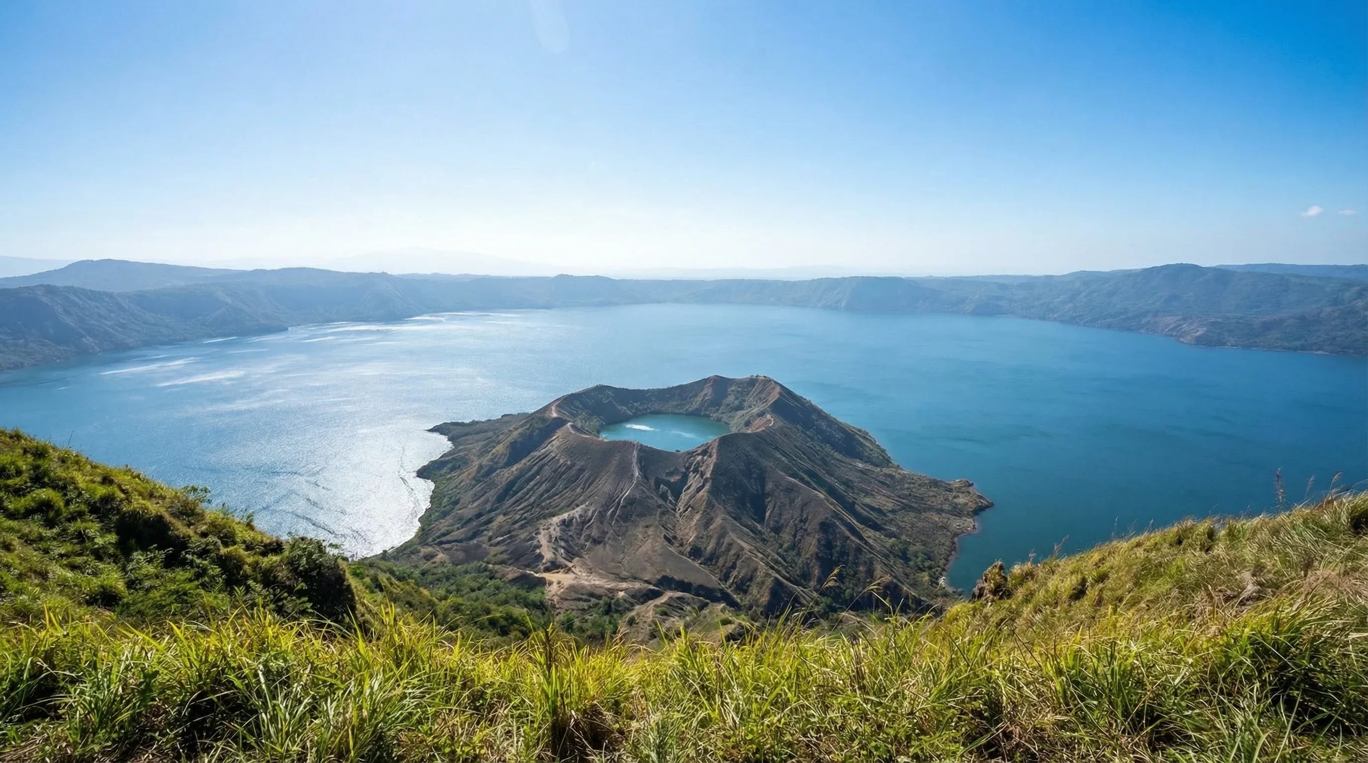 Wide-angle view of Taal Volcano island situated within a large lake, viewed from an elevated ridge under a clear sky.