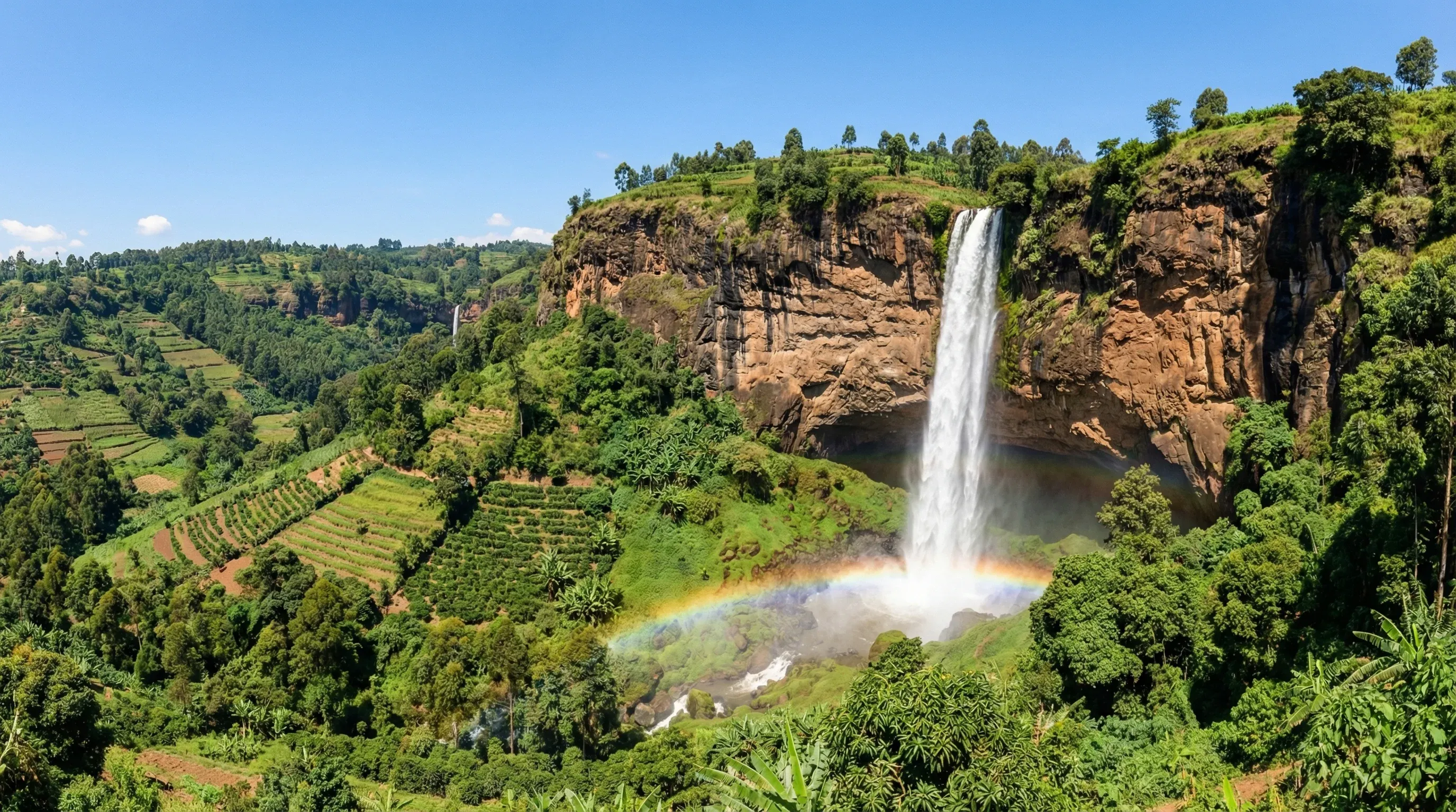 A large waterfall cascading over a steep cliff edge into a green valley under a clear sky at Sipi Falls, Uganda.