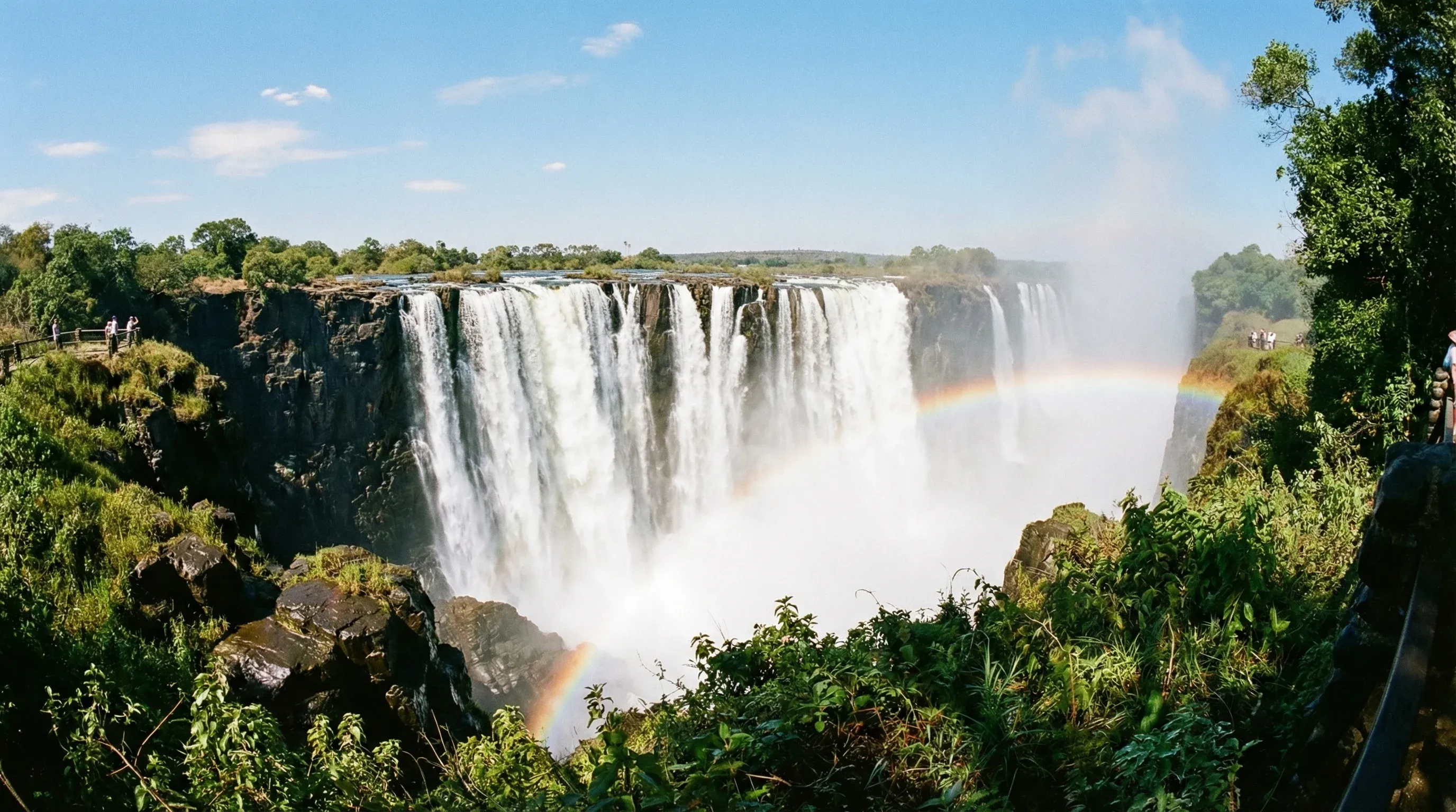 A wide view of the waterfall at Victoria Falls with mist rising and a rainbow visible in the spray.