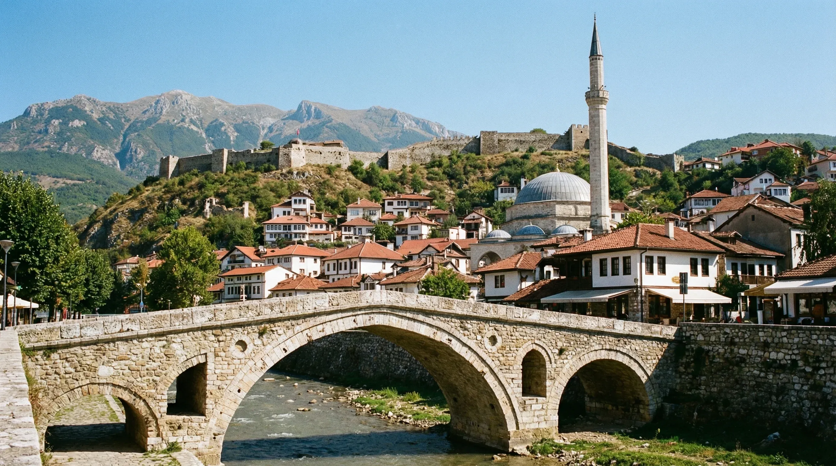 A view of the historic Stone Bridge in Prizren crossing the Lumbardhi river, with the Sinan Pasha Mosque and the hilltop fortress in the background.